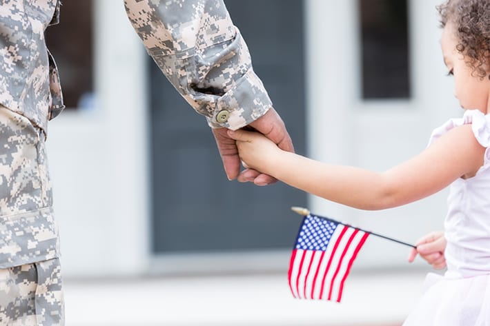 Child in white dress with a small American flag holds hand of military service-member in uniform